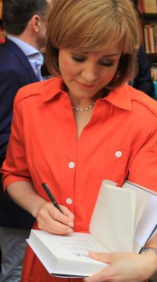 Sian in an orange shirt signs a book with a pen. Bookshelves in the background. The setting is calm and focused.