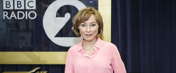 Sian in a pink blouse stands in front of a BBC Radio 2 sign. Dark blue curtain backdrop, bright lighting, calm expression.