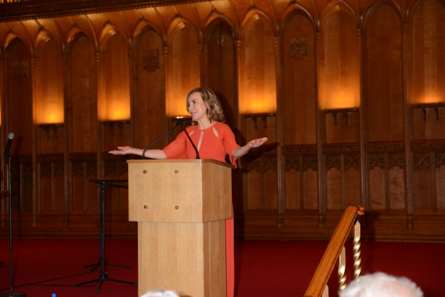 Sian Wlliams in an orange dress speaks at a wooden podium in an ornate hall with soft lighting. Her arms are open in a welcoming gesture.