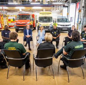 A group of people sit in a circle having a discussion in a garage with emergency vehicles in the background. The mood is attentive.