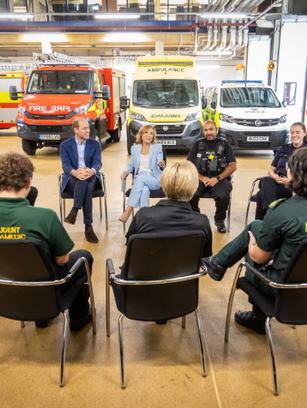 A group of people sit in a circle having a discussion in a garage with emergency vehicles in the background. The mood is attentive.