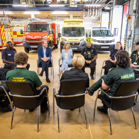 A group of people sit in a circle having a discussion in a garage with emergency vehicles in the background. The mood is attentive.