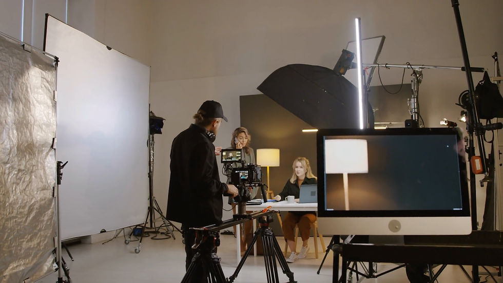 Film crew in studio setting with lighting and camera equipment. A director films a seated person with laptop, reflecting on a monitor.