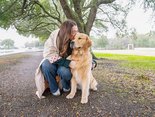 Woman kneels to hug and kiss a golden retriever under a tree on a park path. Overcast sky, lush green grass, and a joyful mood.