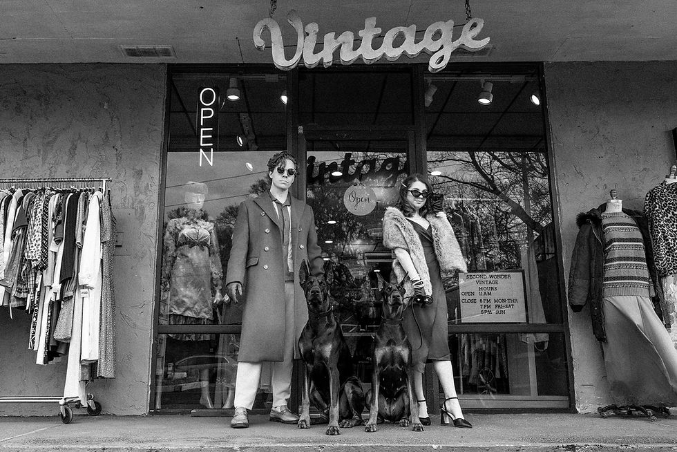 Two stylish individuals with two dogs stand outside a vintage store. Neon "Open" sign and racks of clothes in view. Black and white.