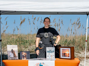Smiling person under a white tent at the beach, behind an orange table with pet photos and "The Photo District" sign. Blue sky, seagrass in back.