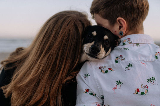 Two people cuddle a small black dog by the water at dusk, one in a palm-tree shirt, creating a tender mood.
