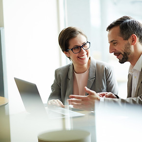 Two professionals smiling and collaborating at a desk while reviewing information on a laptop in a bright office setting.