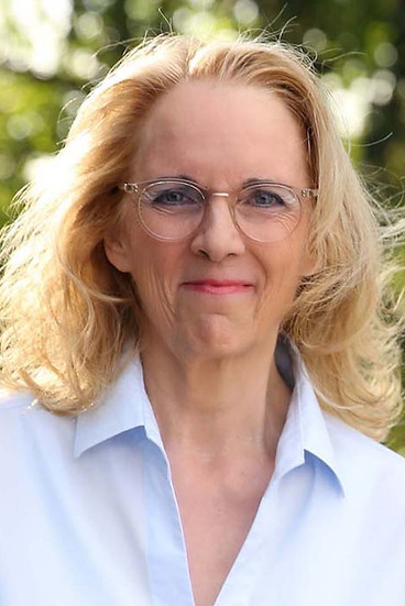 Porträt von Martina Schwanke in Nahaufnahme, mit sanftem Lächeln und grünem Waldhintergrund voller natürlicher Lichtreflexionen.