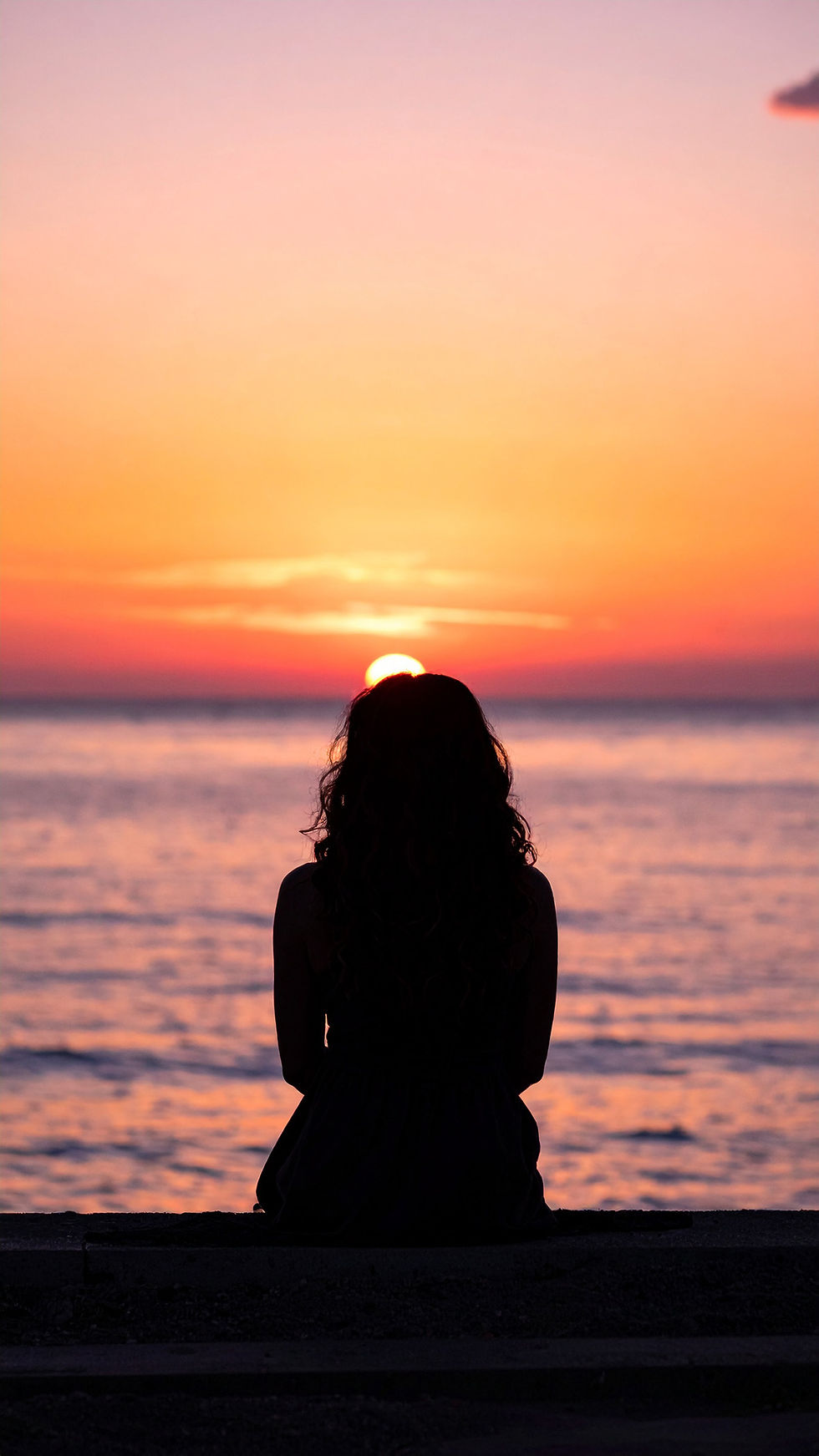 A lady contemplating on the beach, she's facing the horizon as the sun is setting.