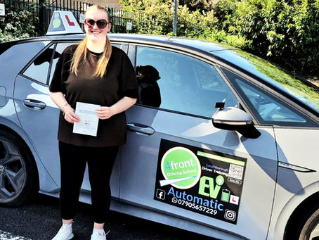 A smiling Stephanie holding up her first-time pass driving test certificate outside the Loughborough Driving Test Centre, standing in front of a white 4front Driving School electric VW ID3 car.