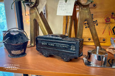 A black metal toy train car sits on a shelf. Gold lettering reads "New York Central". A stamp dispenser, and mini black bucket sit on either side.