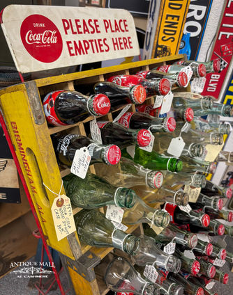 Old yellow and red bottle crates on display containing a variety of empty and full coke bottles. A sign above reads "Please place empties here".