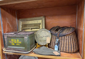 Fishing basket, post office flap, ammo box, a hat, and a large metal seal on a shelf.