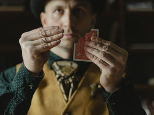 Magicien en gilet et chapeau présentant une carte à dos rouge dans une ambiance feutrée.