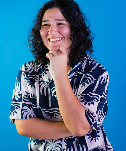 Pessoa sorrindo, com cabelo cacheado, sentada em frente a um fundo azul, apoiando o queixo com a mão e vestindo roupa estampada em tons de azul e branco.