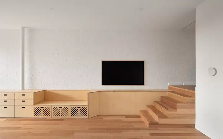 A minimalist living room with light wood floors and a white exposed brick wall. A black television screen is mounted on the wall. Below the screen, a custom-built, light wood cabinet with drawers and open shelves forms a long entertainment unit that transitions into a low bench and built-in storage.