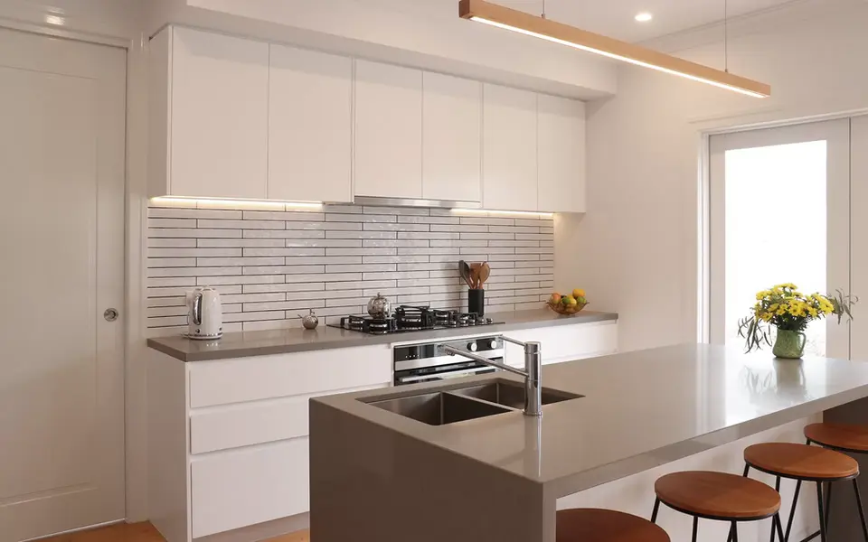 A modern kitchen featuring a gray island with a sink and four wooden-topped bar stools. The main kitchen wall has a white subway tile backsplash, white upper cabinets, and a stove and range hood. A long, wooden light fixture hangs above the island. On the counter, a vase of yellow flowers adds a pop of color.