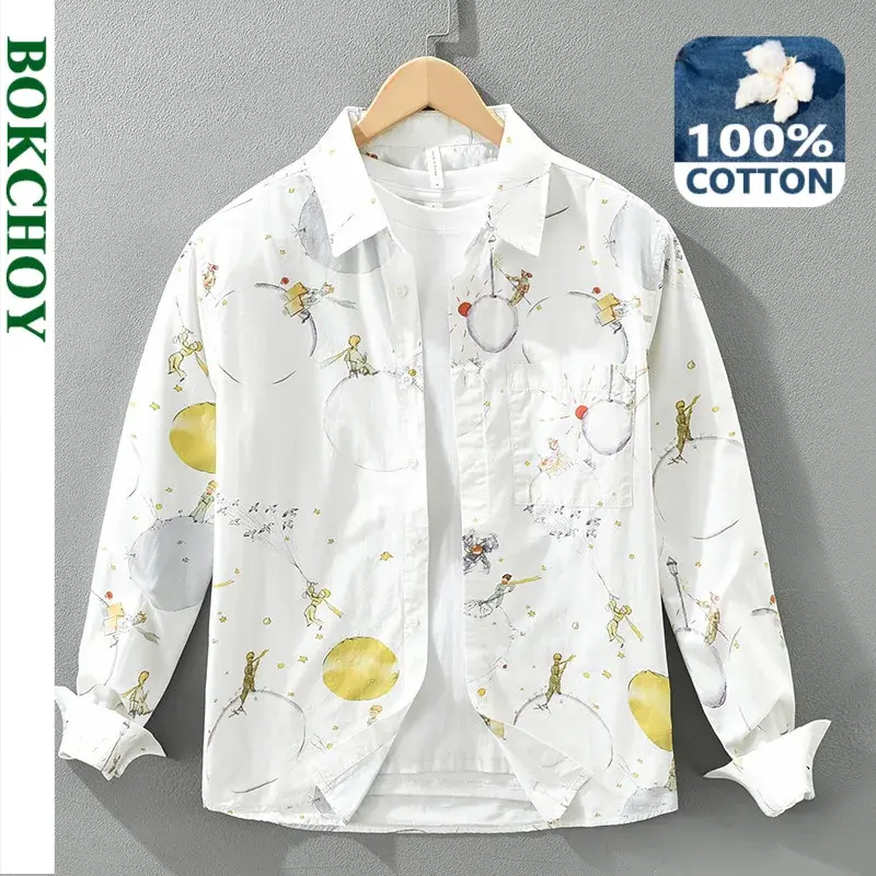 White shirt with whimsical illustrations of small figures and planets, hung on a wooden hanger. 100% cotton label visible.