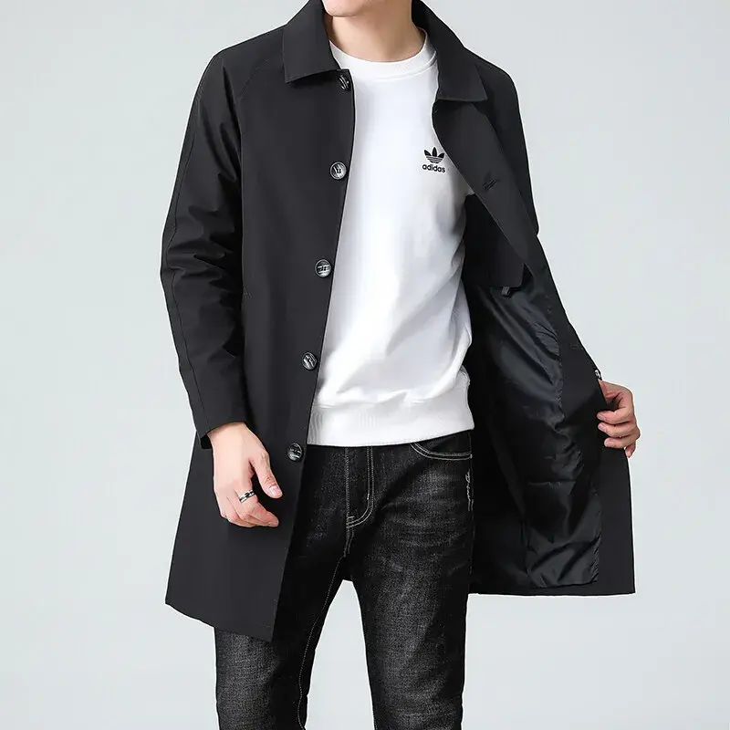 Man wearing a black coat, white shirt, and dark jeans, holding the coat open, against a light gray background.