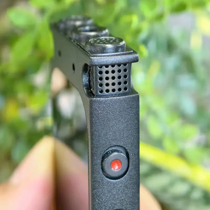 Thumbnail: Close-up of a black perforated object with a red button, set against a blurred green leafy background.