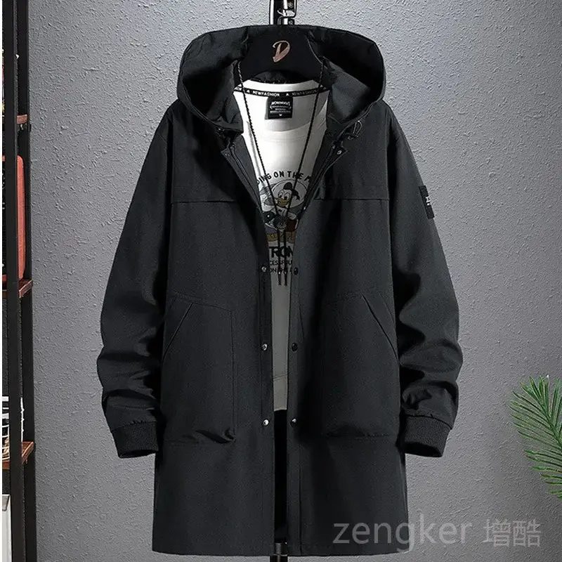 Black hooded coat on hanger over printed white shirt, against gray textured wall with a green plant on the lower right.