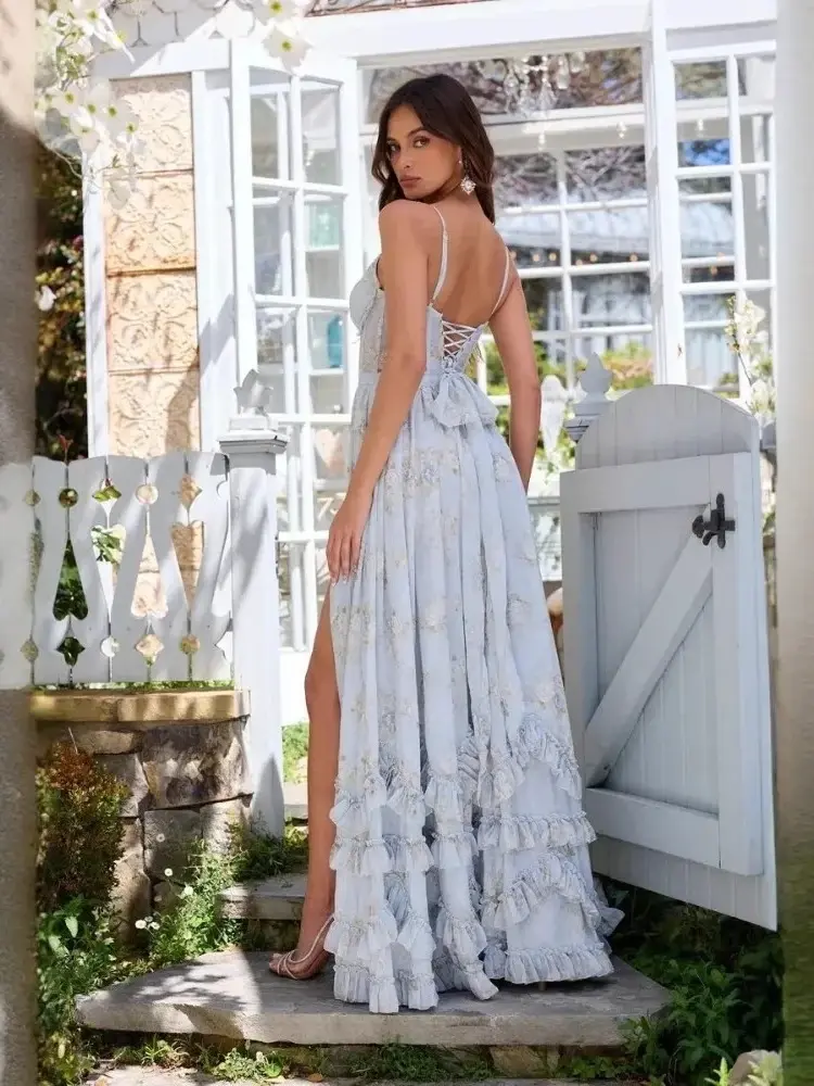 Pikkukuva: Woman in a flowing blue dress with lace-up back stands in a garden by a white gate, looking over her shoulder.