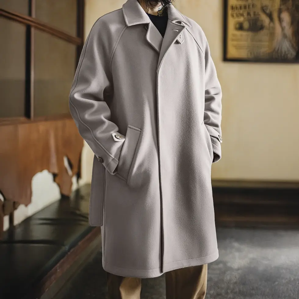 Man in a light gray coat with hands in pockets, standing indoors with a rustic interior background.