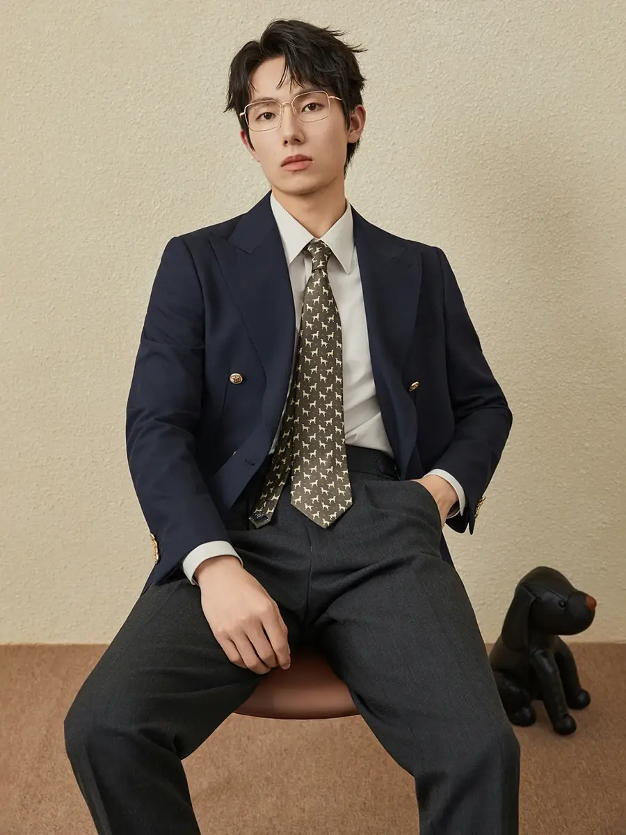 Thumbnail: Man in a navy blazer, white shirt, patterned tie, and gray trousers, stands against a beige background.