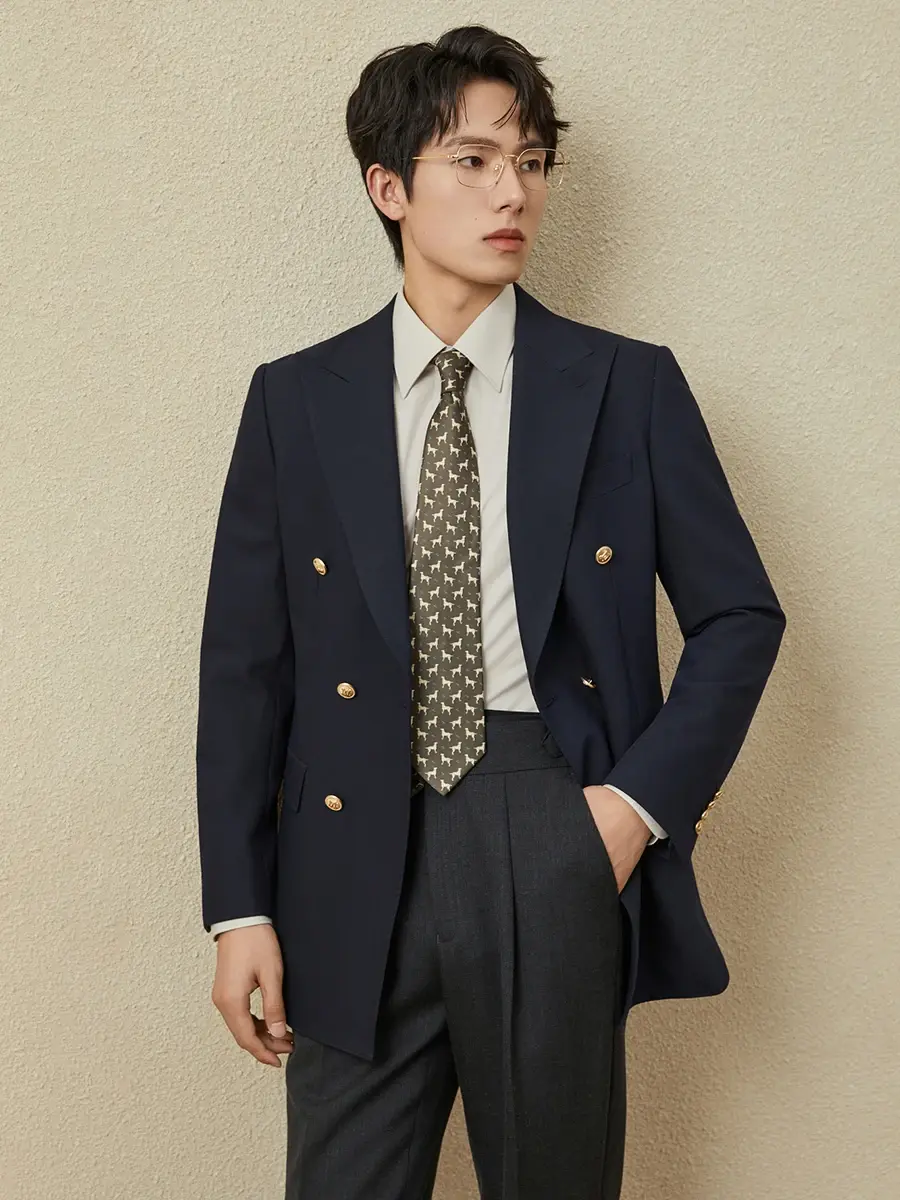 Thumbnail: Man in a navy blazer, white shirt, patterned tie, and gray trousers, stands against a beige background.