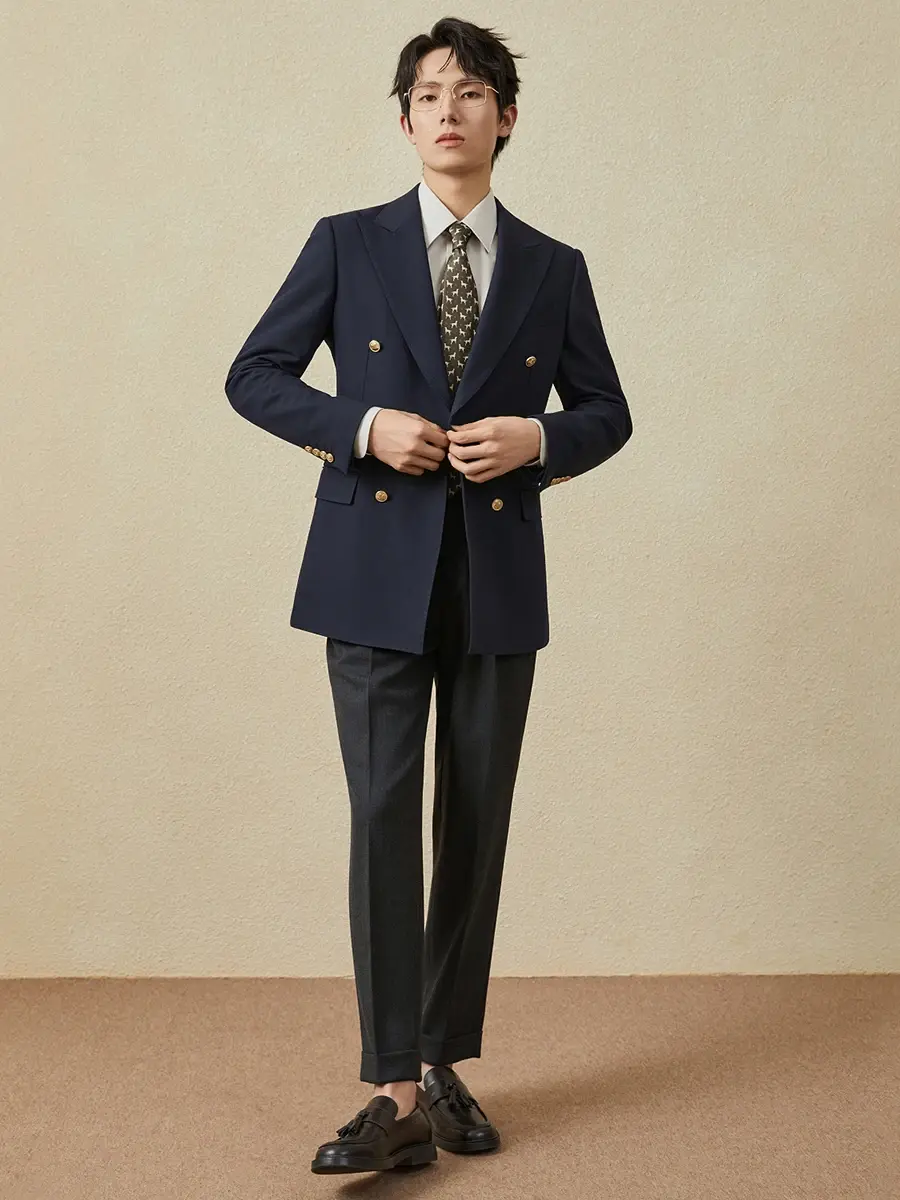 Thumbnail: Man in a navy blazer, white shirt, patterned tie, and gray trousers, stands against a beige background.