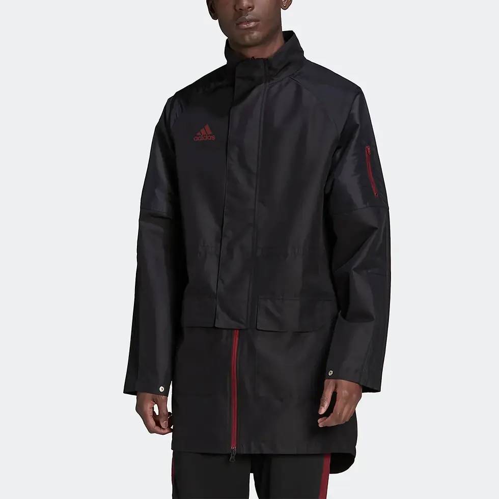 Miniatura: Man in a long black jacket with red details, standing against a gray background.