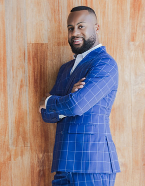 Picture of Luzerne with body turned to the right and facing forward. He is smiling. His arms are folded. He is wearing a blue with white pin-checkered suit over a white, patterned shirt. He stands in front of light brown wooden wall.
