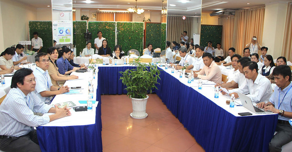 Formal meeting in a hotel conference room, people seated around U-shaped tables with laptops and papers, a potted plant in center.