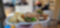 A close-up of a plate featuring a toasted sandwich with lettuce and a pickle on top, served with a side of potato chips. The background shows the blurred interior of the coffeehouse.