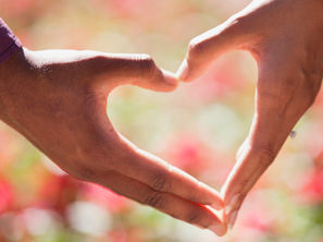 Two hands forming a heart shape against a blurred pink and green background, evoking feelings of love and connection.