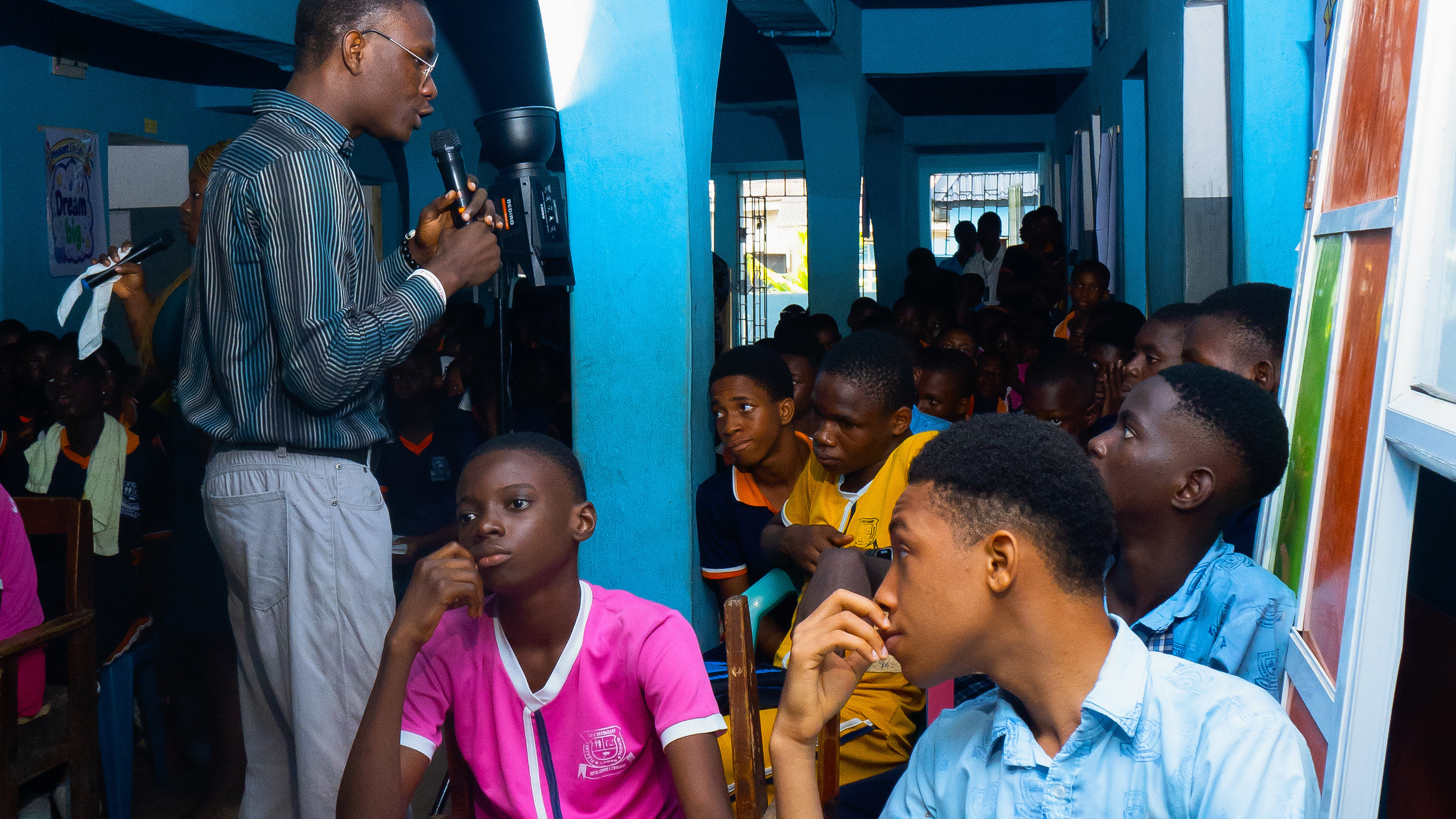A male facilitator wearing glasses and a striped shirt speaks into a microphone during an interactive school outreach session. Students in colourful uniforms sit attentively in a blue-walled hall, listening to the presentation and participating in the Africa 10XG Ethical Leadership programme.