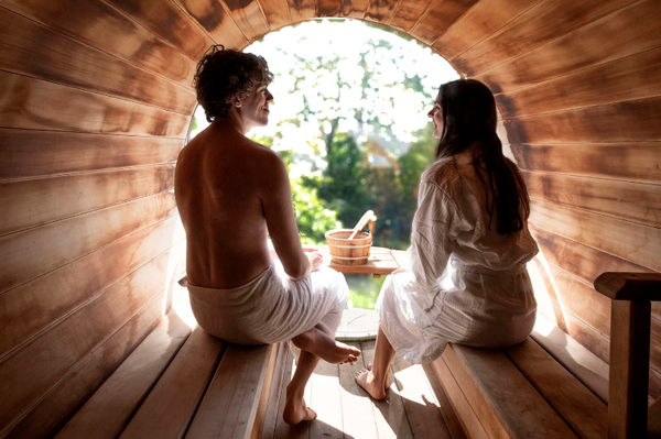 Deux personnes en peignoir dans un sauna en bois rond, discutant devant une fenêtre avec vue sur un jardin verdoyant. Atmosphère paisible.