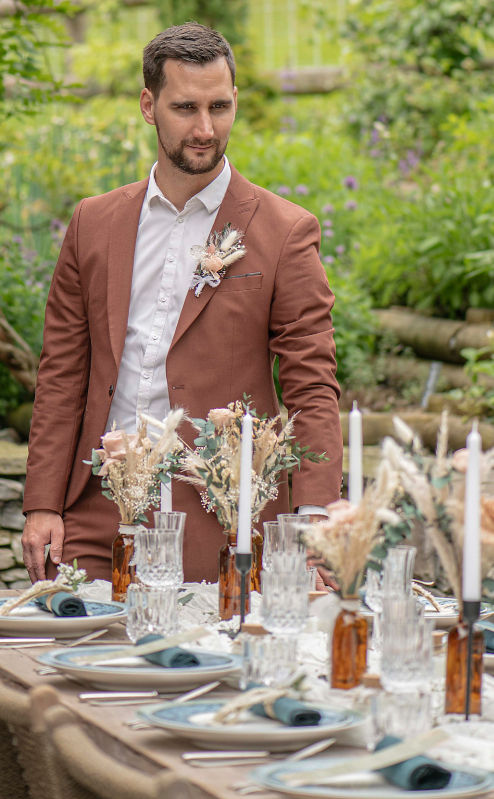Un homme en costume beige avec fleur à la boutonnière se tient près d'une table élégante décorée de bougies et fleurs, extérieur verdoyant.