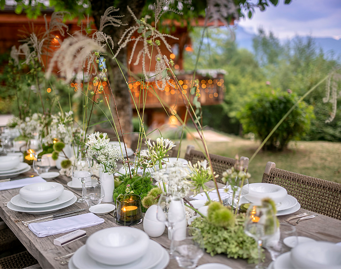 Une élégante table dressée avec des compositions florales et de la porclaine blanche dans le jardin d'un chalet de montagne.