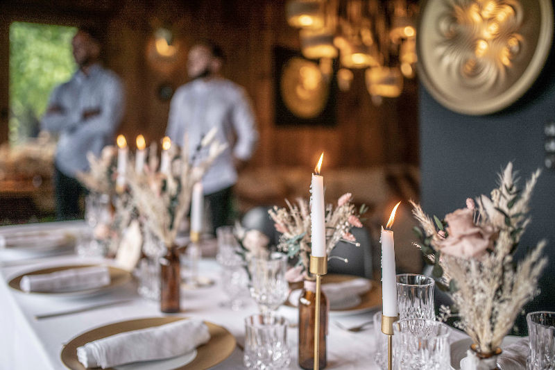 Table élégamment dressée avec chandeliers et fleurs séchées. Deux hommes flous en arrière-plan. Ambiance chaleureuse et boisée.