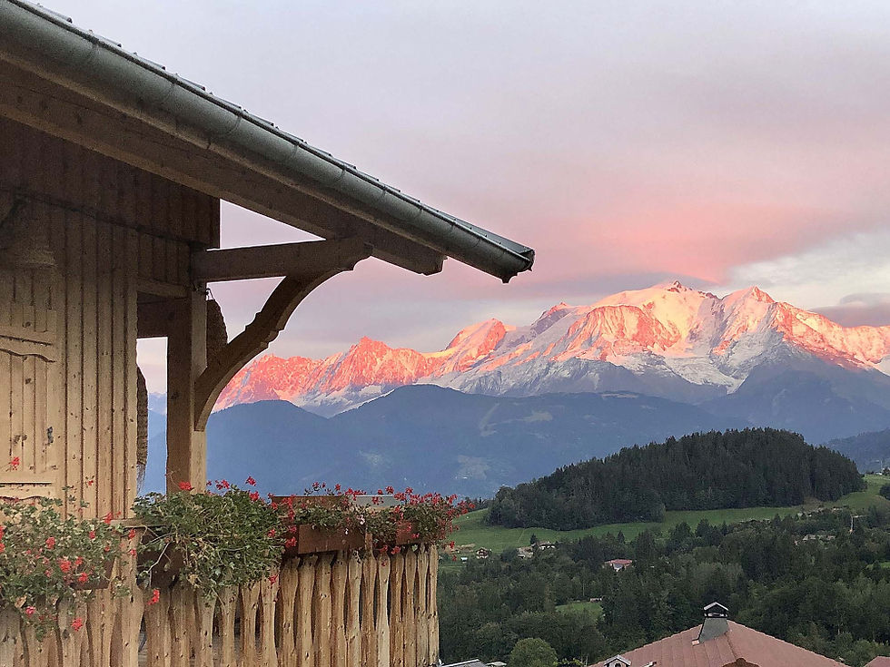 Balcon de chalet fleuri au premier plan, face à des montagnes enneigées roses au coucher du soleil, dans une vallée verdoyante.