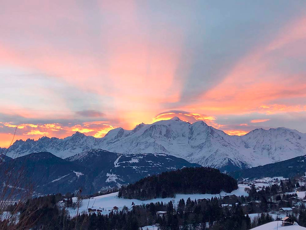 Chaîne de montagnes enneigées au lever du soleil, ciel rose et orange au-dessus d’un village alpin et de forêts.