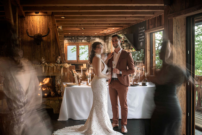 Mariés souriants dans un chalet rustique, devant un buffet et une cheminée, avec invités flous en mouvement.