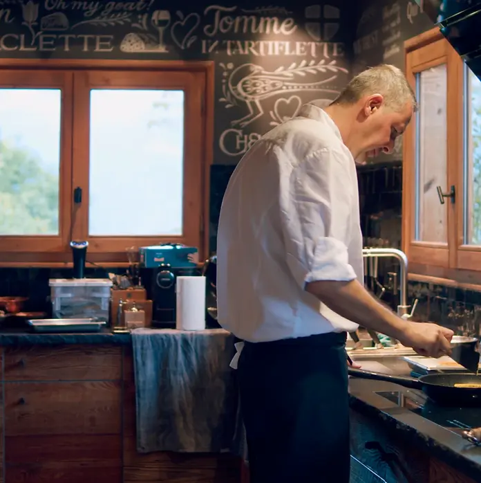 Chef en veste blanche cuisinant dans une cuisine rustique, devant fenêtres en bois et tableau noir avec RACLETTE, TARTIFLETTE.