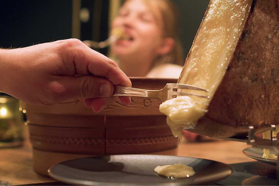 Main servant du fromage fondu à la raclette dans une assiette, avec un enfant flou en arrière-plan.