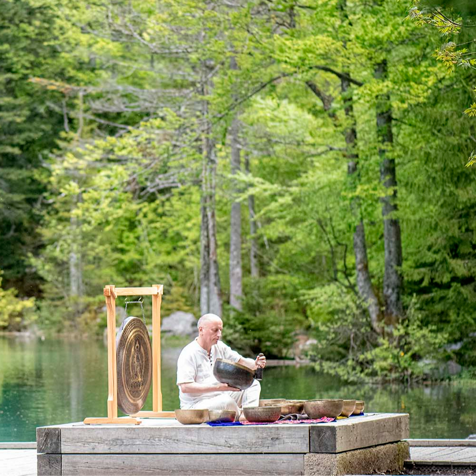 Un homme en blanc joue des bols chantants et un gong près d'un lac entouré d'arbres verts, créant une ambiance paisible et sereine.