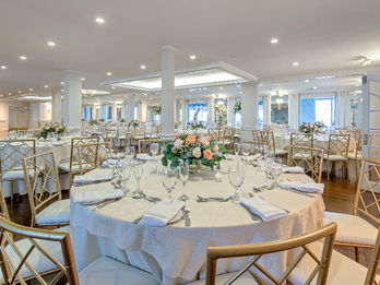 Close-up of a decorated banquet table with floral centerpiece, folded napkins, glassware, and gold chairs in a bright hall.