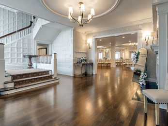 Elegant venue entrance with a chandelier, wood flooring, white staircase, and view into a formal dining room beyond.