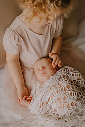 Baby broertje bij grote zus op de schoot tijdens een newborn lifestyle fotoshoot in Mechelen door Souvenirs de Zoë Fotografie. Grote zus aait verterend haar klein broertje.
